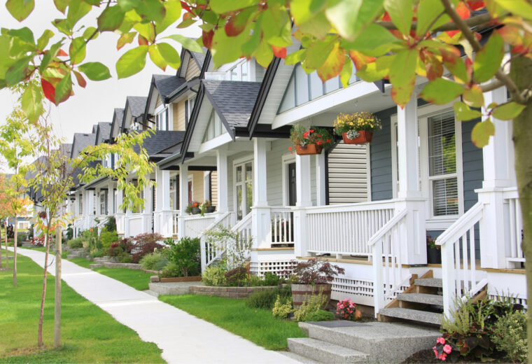 A row of identical townhouses with home insurance small front yards and sidewalks, under a canopy of green and red leaves.